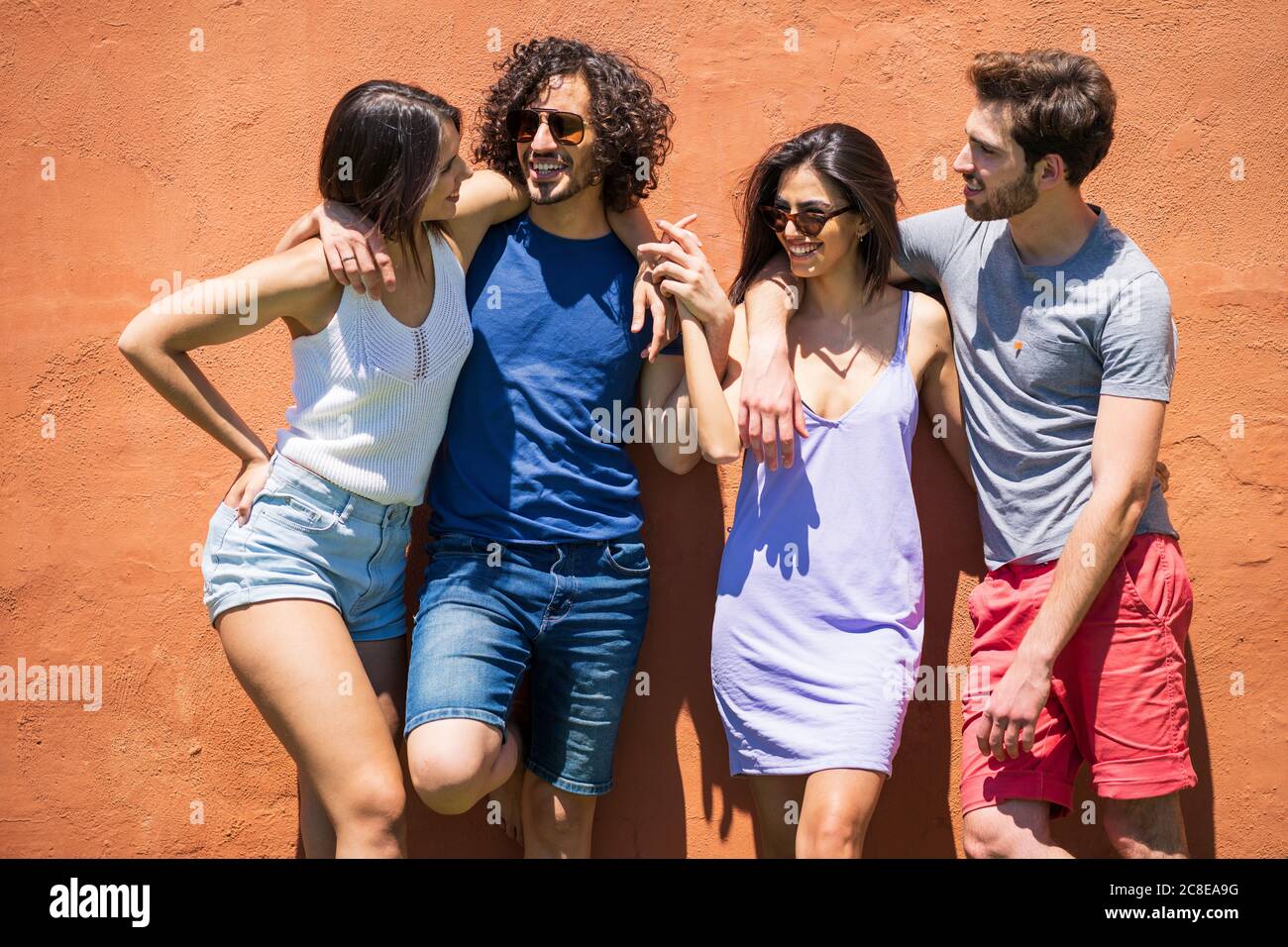 Felici giovani amici multietnici maschili e femminili in piedi contro il marrone muro al cortile posteriore in giornata di sole Foto Stock