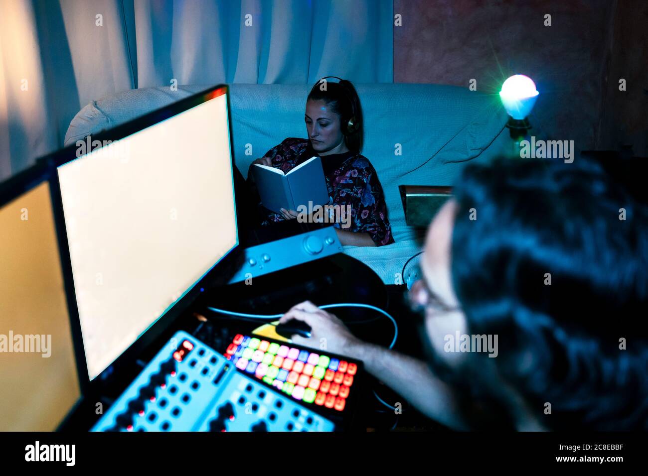 Uomo che usa il computer mentre donna legge il libro in studio di registrazione a casa Foto Stock
