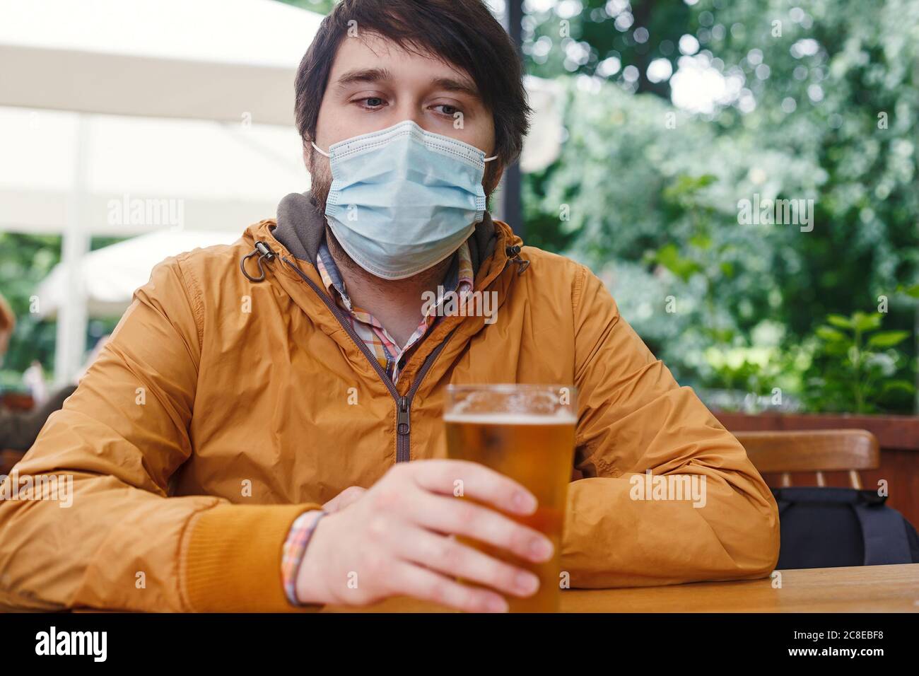 Uomo caucasico con bicchiere di birra seduto in una maschera medica sulla veranda del caffè. Nuovo concetto normale Foto Stock