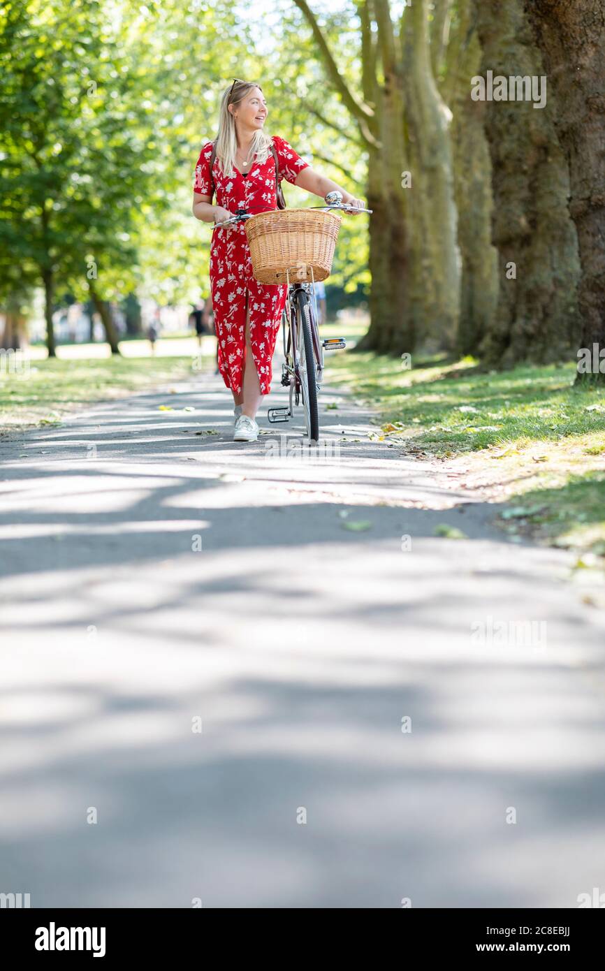 Donna che guarda via mentre cammina con la bicicletta sul sentiero dentro parco pubblico durante il giorno di sole Foto Stock