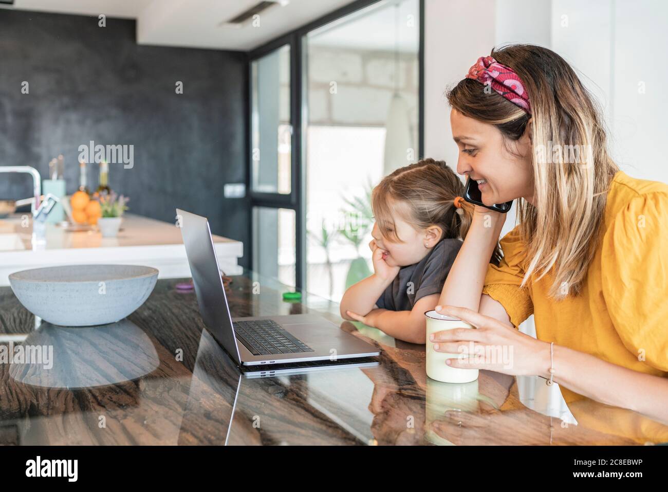 Madre con figlia che guarda il notebook mentre parla con intelligenza telefono in casa Foto Stock