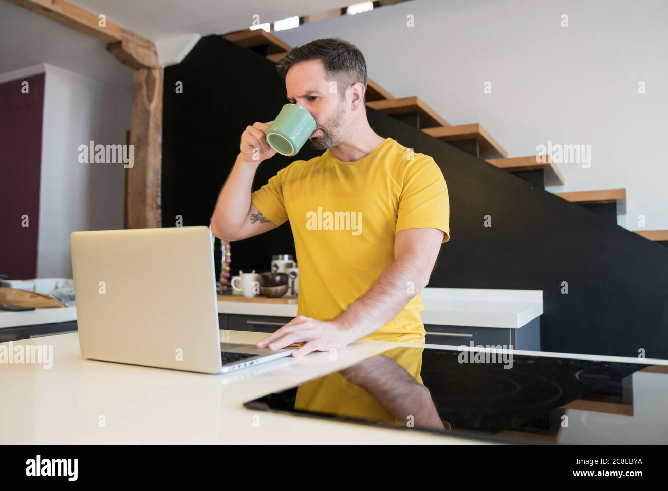 Uomo che beve il caffè mentre usa il computer portatile sull'isola della cucina a. casa Foto Stock