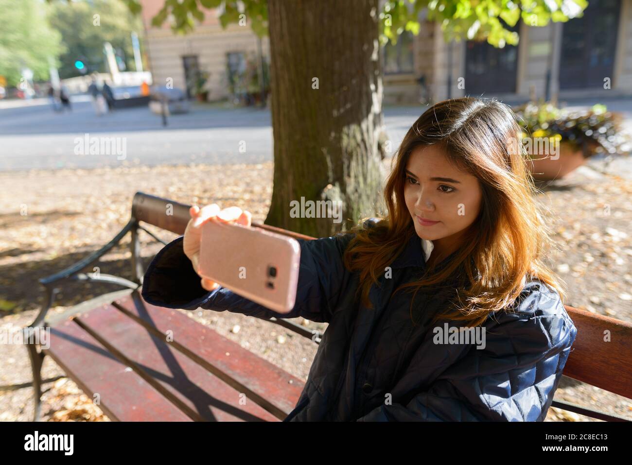 Giovane bella donna asiatica che prende selfie con il telefono cellulare seduto su panca sotto l'albero verde alto Foto Stock