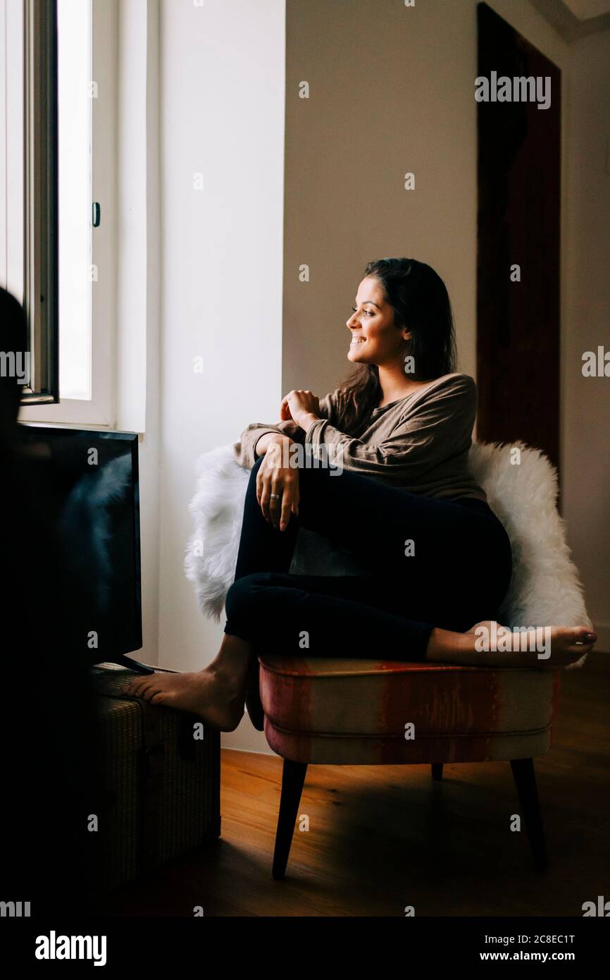 Giovane donna sorridente che guarda attraverso la finestra mentre si siede sulla sedia a casa Foto Stock