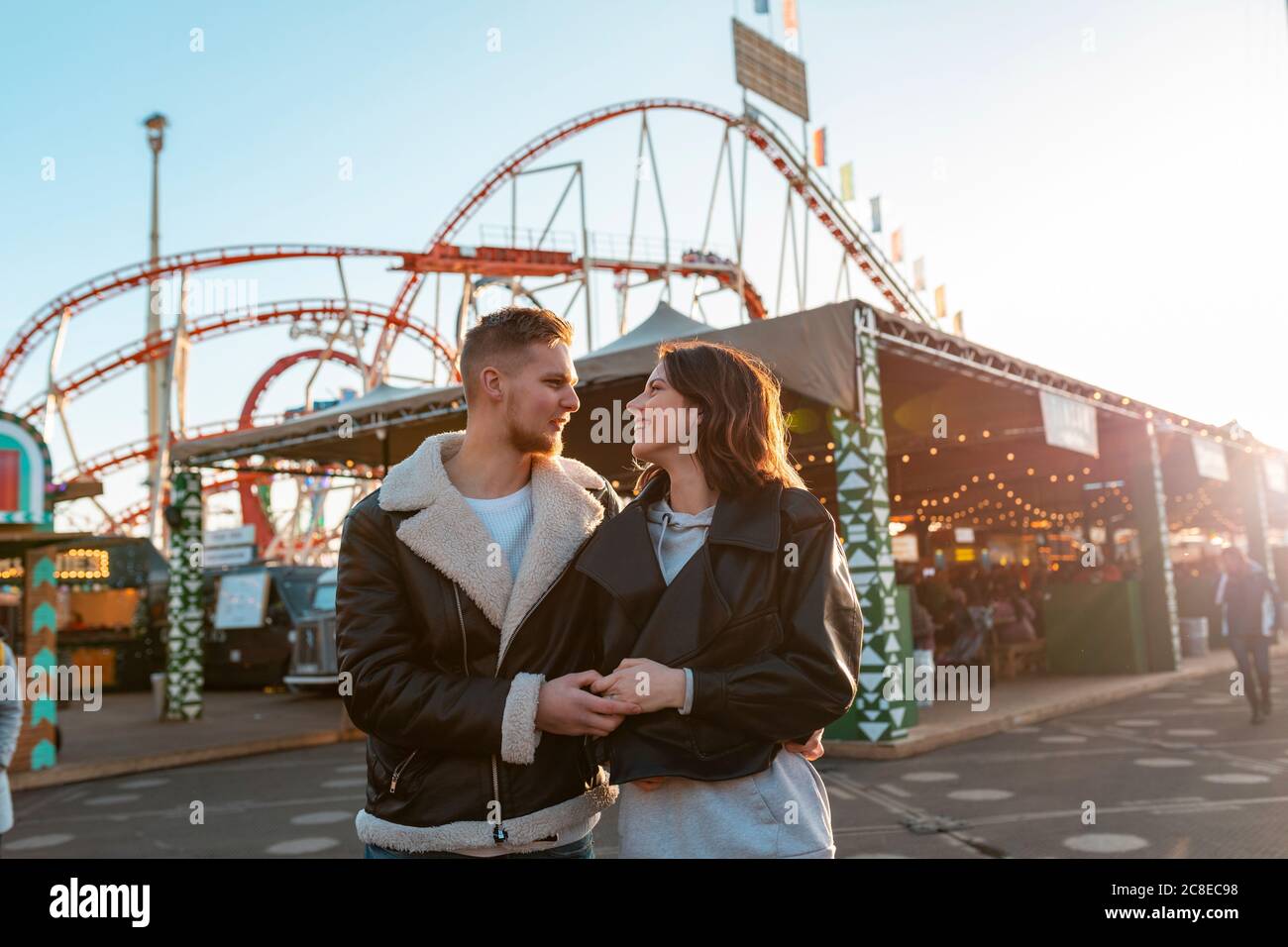 Giovane coppia romantica che tiene le mani mentre si trova nel parco divertimenti durante il tramonto Foto Stock