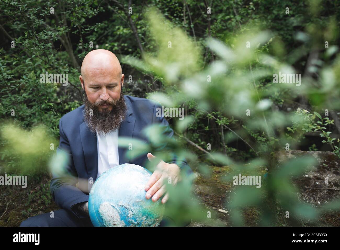 Uomo d'affari calvo che tiene il globo mentre si siede nella foresta Foto Stock