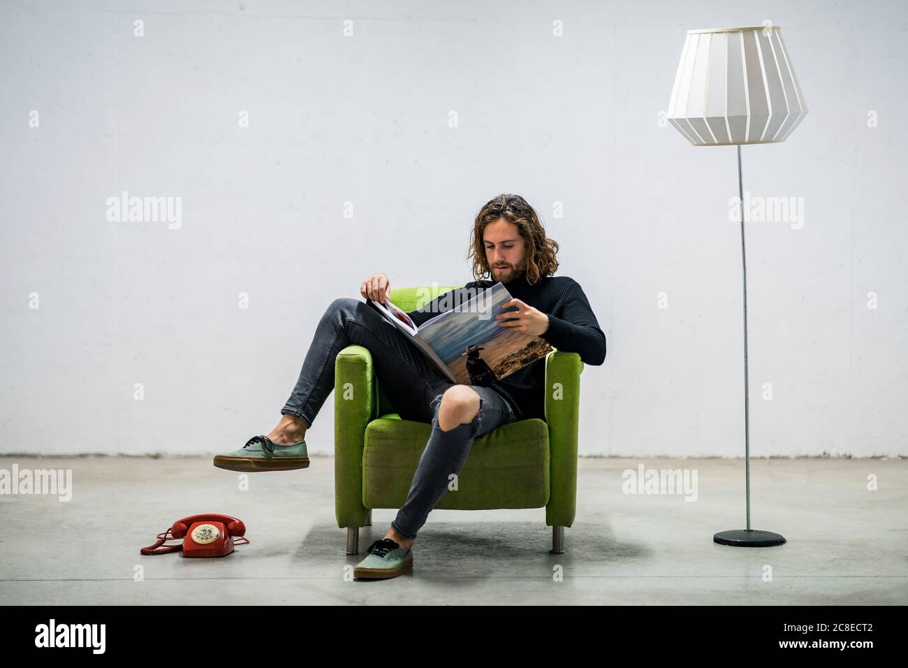 Giovane uomo che legge libro mentre si rilassa sulla poltrona contro il muro a casa Foto Stock