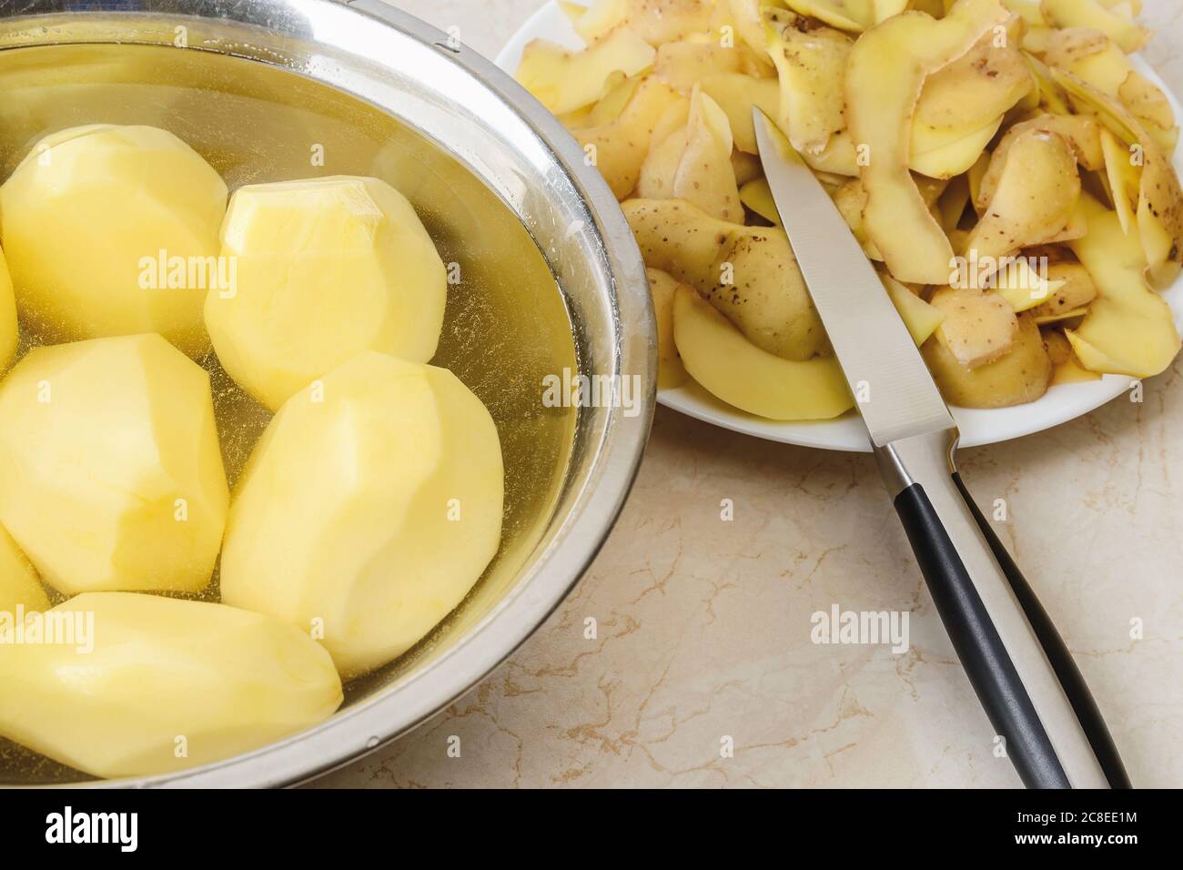 Patate sbucciate grezze in una ciotola con acqua, bucce di patate e un coltello vegetale su un piatto sul tavolo da cucina. Cuocere le patate bollite a casa. Foto Stock