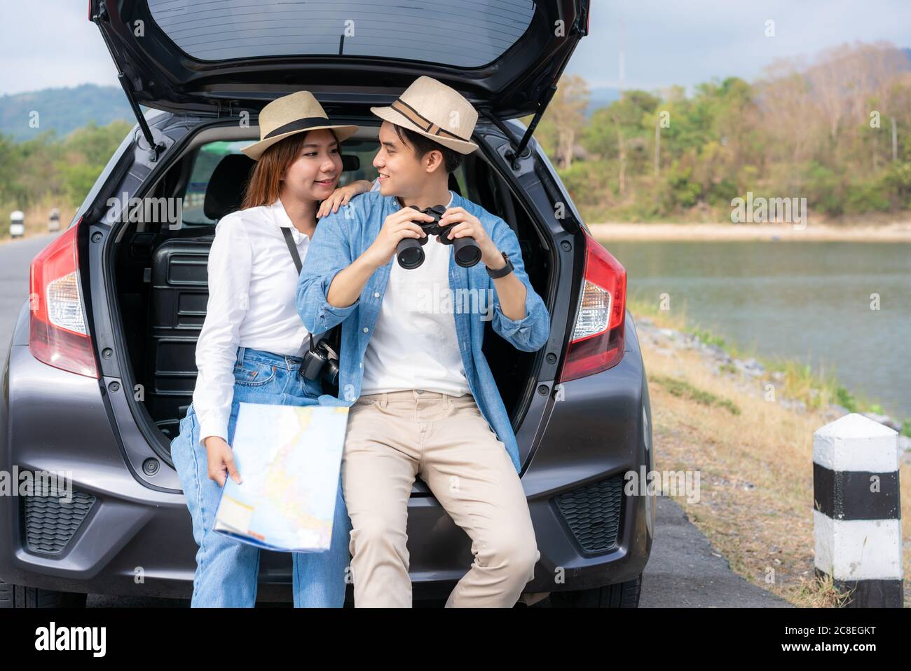 Giovane coppia asiatica con uomo turista che tiene binocoli con ragazza sorridente seduto vicino con mappa su tronco auto in vista campo rurale con lago e m Foto Stock