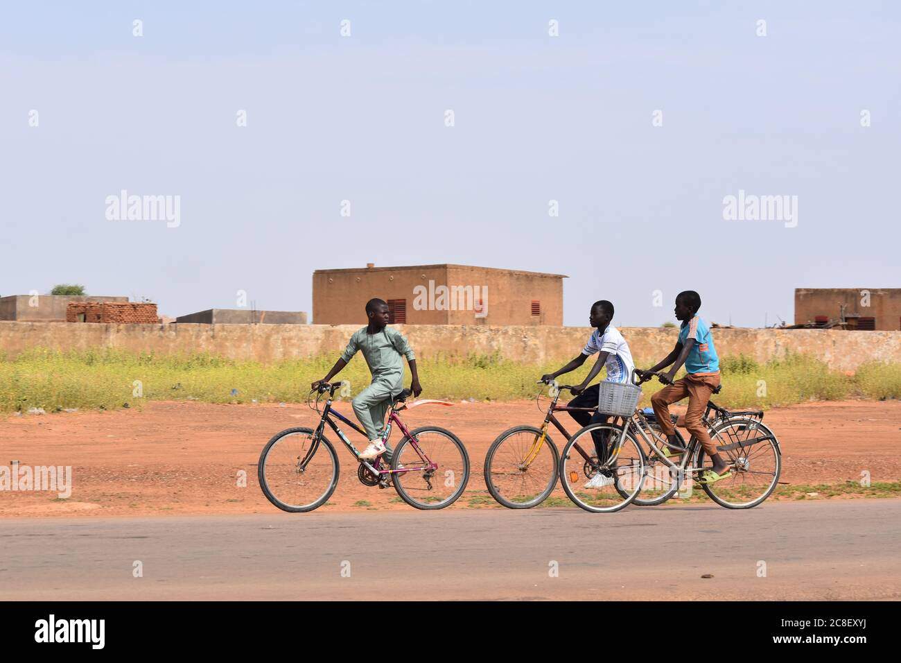 Tre ragazzi adolescenti che cavalcano le biciclette sulla strada principale attraversando un villaggio africano. Foto Stock