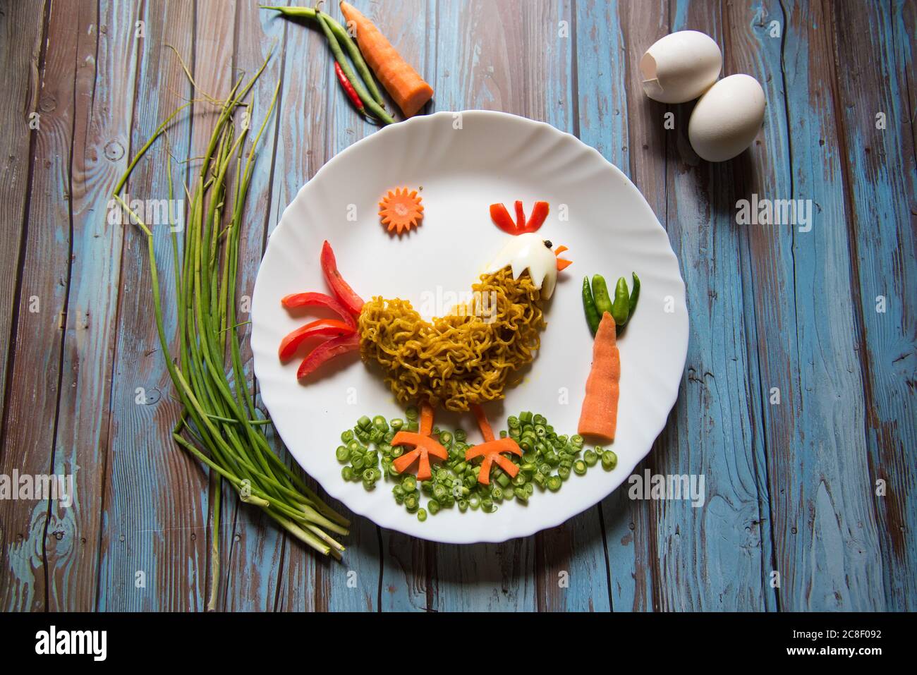 Arte alimentare su sfondo con tagliatelle, verdure, uova sotto forma di gallo su un piatto con uso di fuoco selettivo. Foto Stock