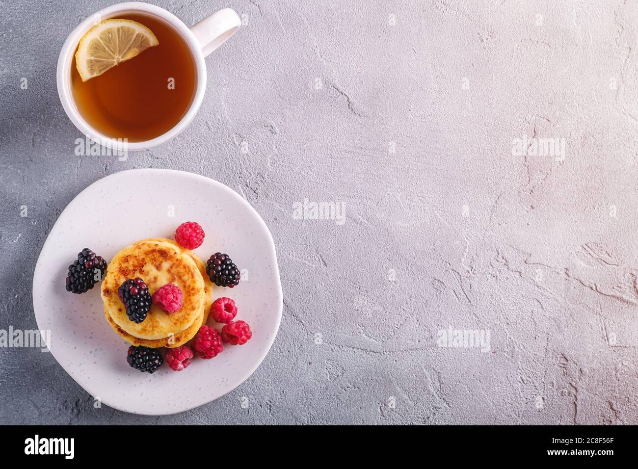 Frittelle di formaggio cottage, frittelle di cagliata dessert con lampone e frutti di bosco in piatto vicino alla tazza di tè caldo con fetta di limone Foto Stock