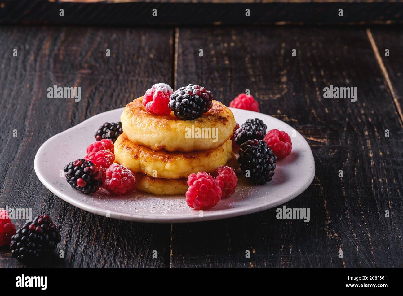 Frittelle di formaggio e zucchero in polvere, frittelle di cagliata dessert con lampone e frutti di bosco in piatto su sfondo nero scuro di legno Foto Stock