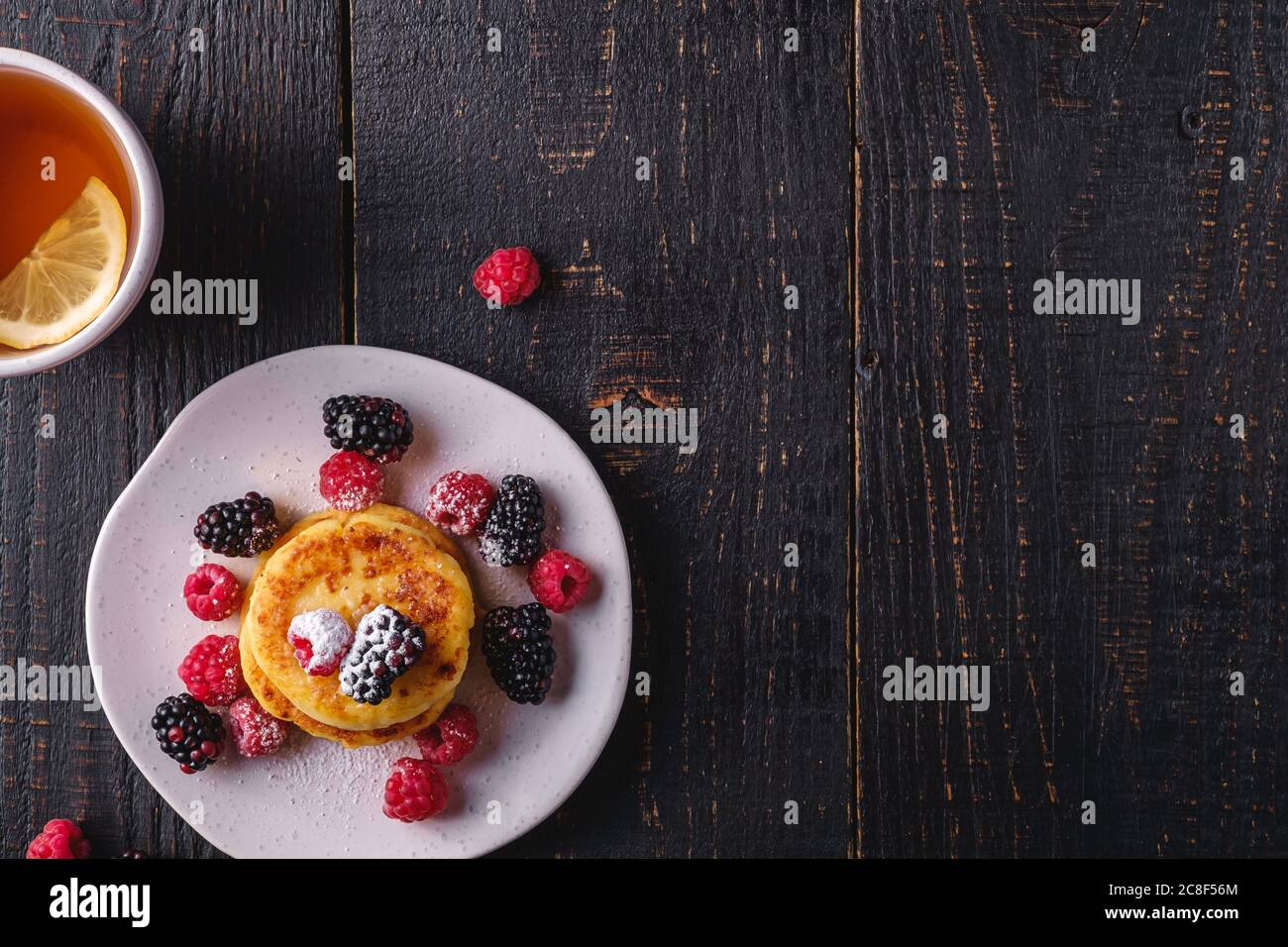 Frittelle di formaggio cottage, frittelle di cagliata dessert con lampone e frutti di bosco in piatto vicino alla tazza di tè caldo con fetta di limone Foto Stock