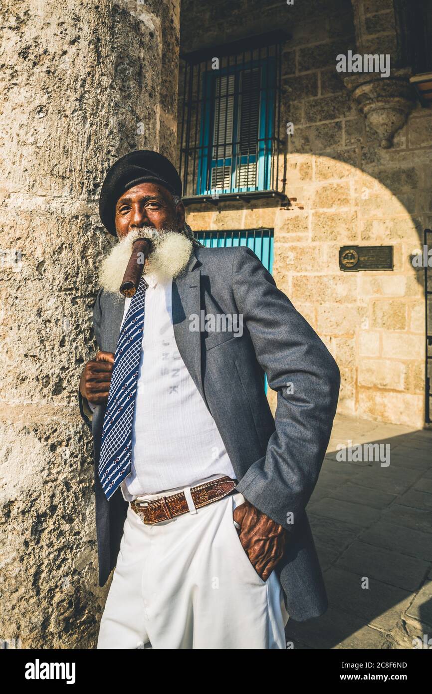 Havana / Cuba - 04.16.2015: Afro cubano vecchio con una lunga barba bianca che indossa un vestito e una berta militare nera, fumando un grande sigaro Foto Stock