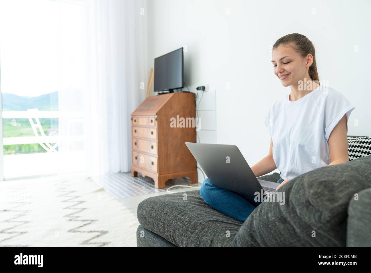 Sorridente giovane donna attraente seduta sul divano usando il laptop che comunica lavorando in linea a casa, ragazza teen felice che scrive sul computer Foto Stock