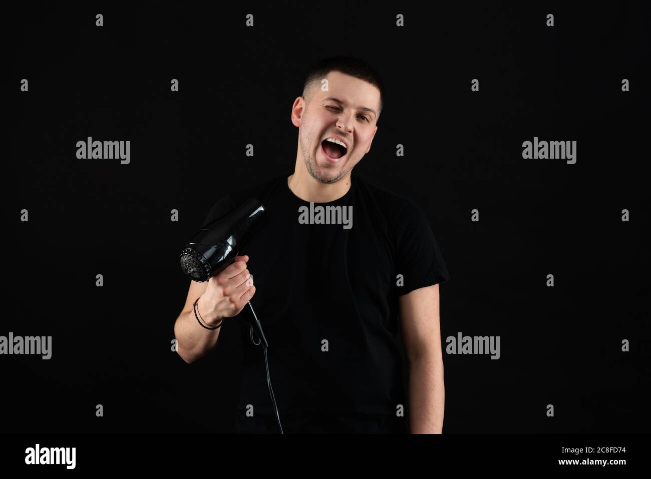 Divertente giovane uomo 20s anni in nero t-shirt tenere soffio capelli asciutti isolato su sfondo nero parete, studio ritratto Foto Stock