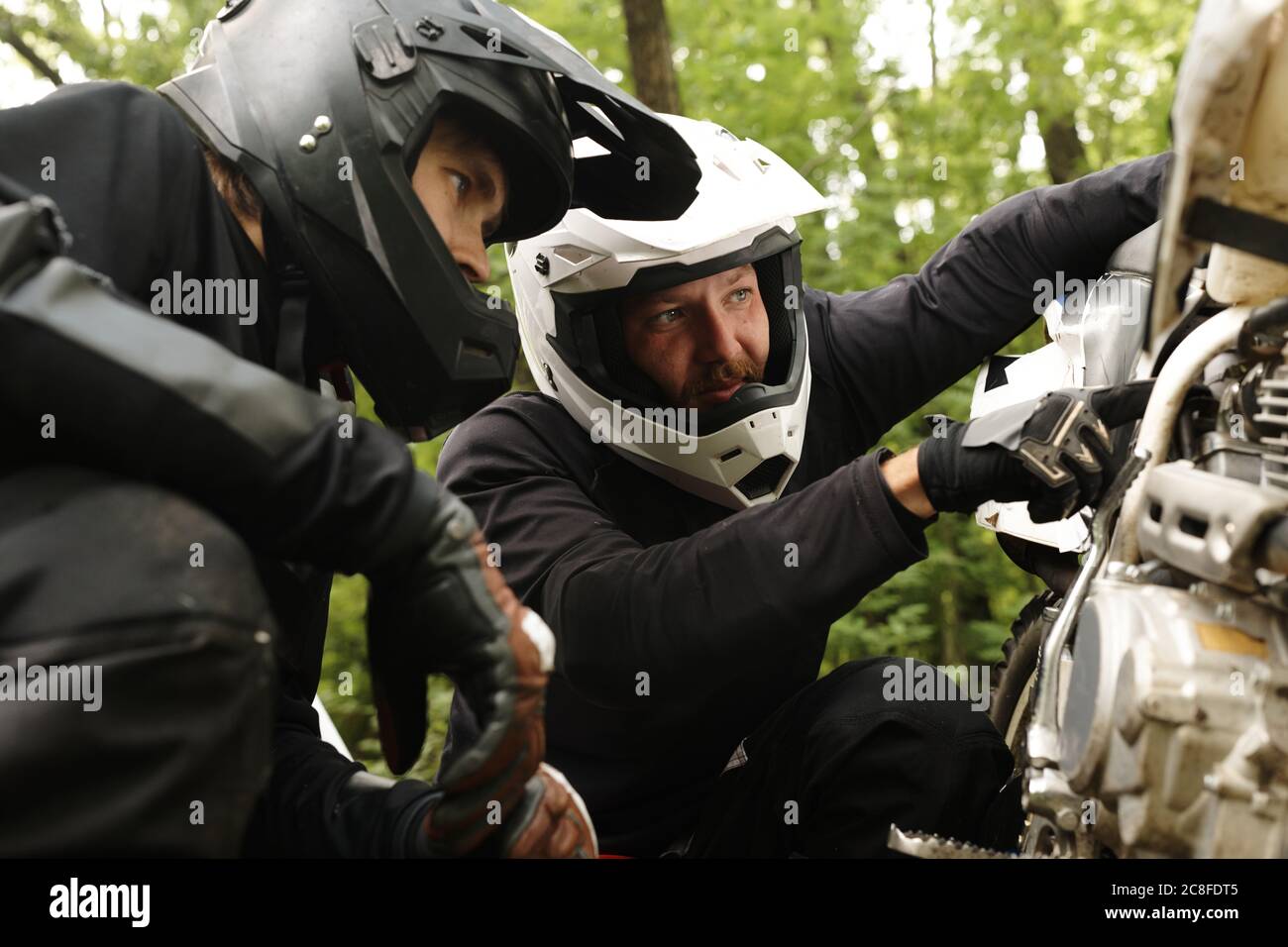 Motociclista concentrato in casco che punta al motore, mentre scoprendo la ragione di guasto moto con amico in foresta Foto Stock
