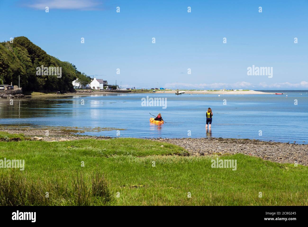 Vista del villaggio con una giovane donna che paddling un kayak in calmo mare blu ad alta marea nella bellissima Red Wharf Bay, Isola di Anglesey, Galles, Regno Unito, Gran Bretagna Foto Stock