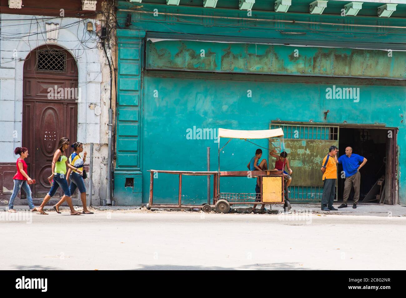 Havana / Cuba - 04.15.2015: Tre donne cubane che camminano di fronte ad un luogo di lavoro dove due uomini cubani stanno in piedi e parlano, due bambini che giocano su un'olla Foto Stock