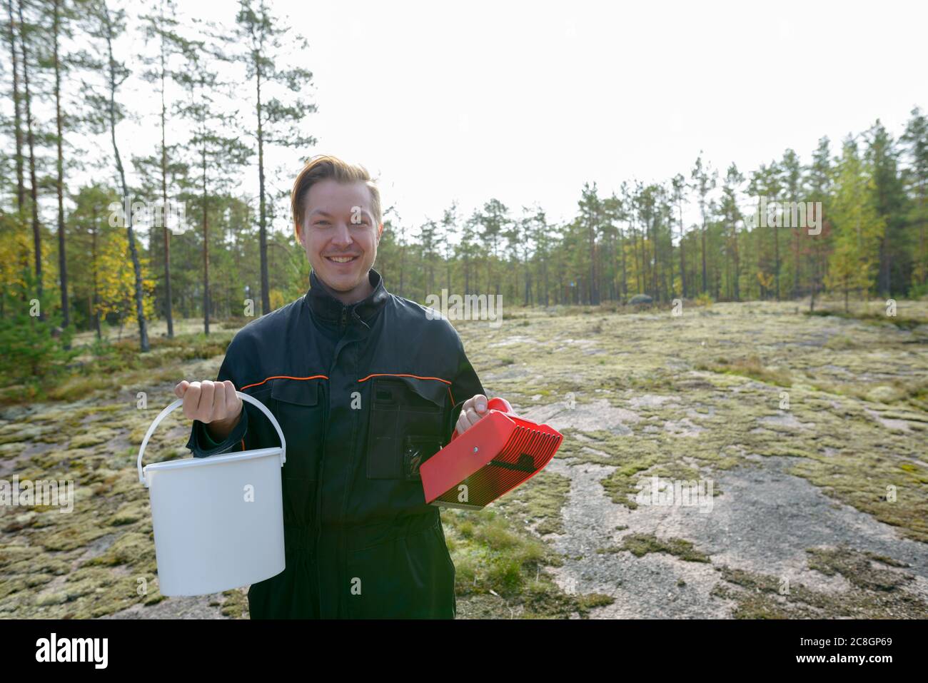 Ritratto di felice giovane bell'uomo che tiene il secchio e picker di bacche nella foresta Foto Stock