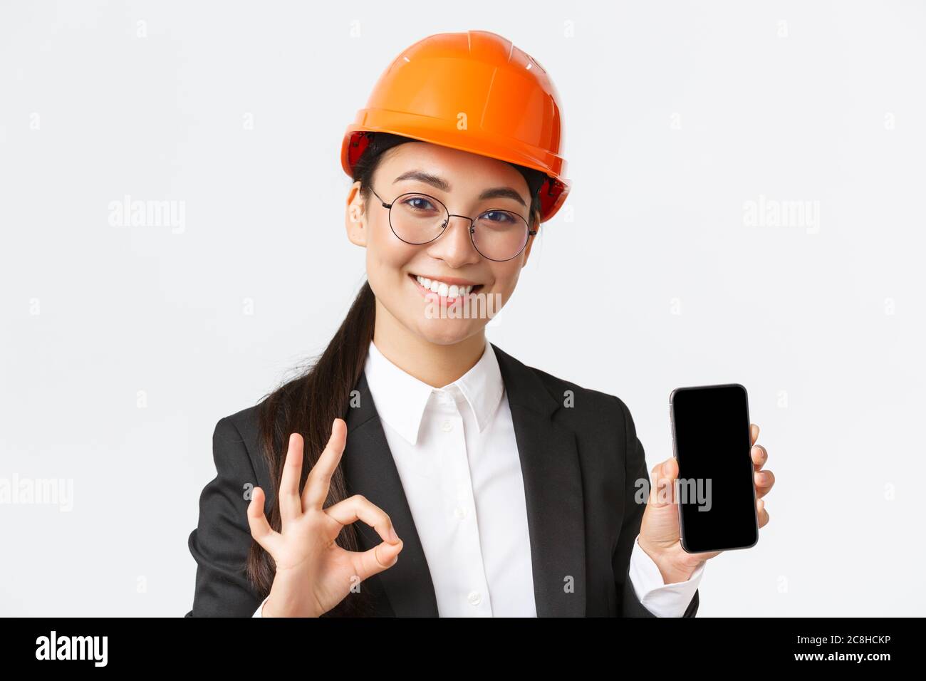 Soddisfatta giovane ingegnere asiatico, architetto in tuta e casco di sicurezza mostrando schermo telefono cellulare e fare un gesto OK, approvare, raccomandare Foto Stock