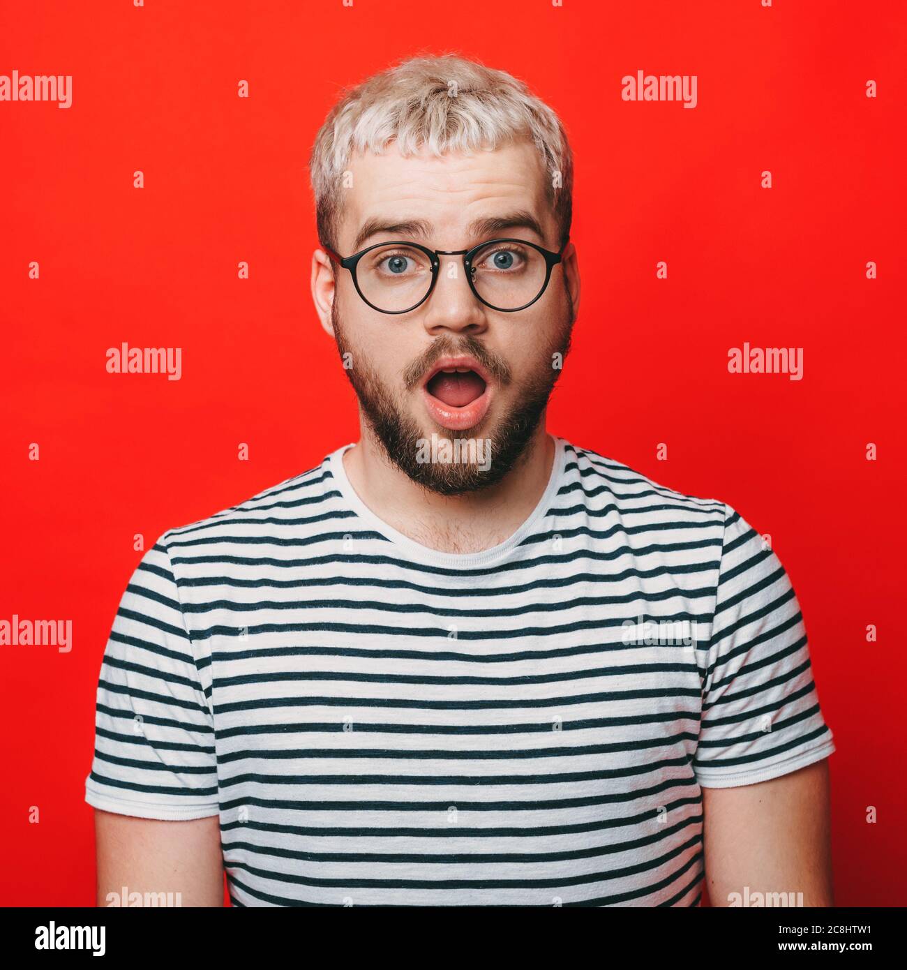 Uomo caucasico biondo gesturing stupore mentre indossa occhiali su una parete rossa studio Foto Stock