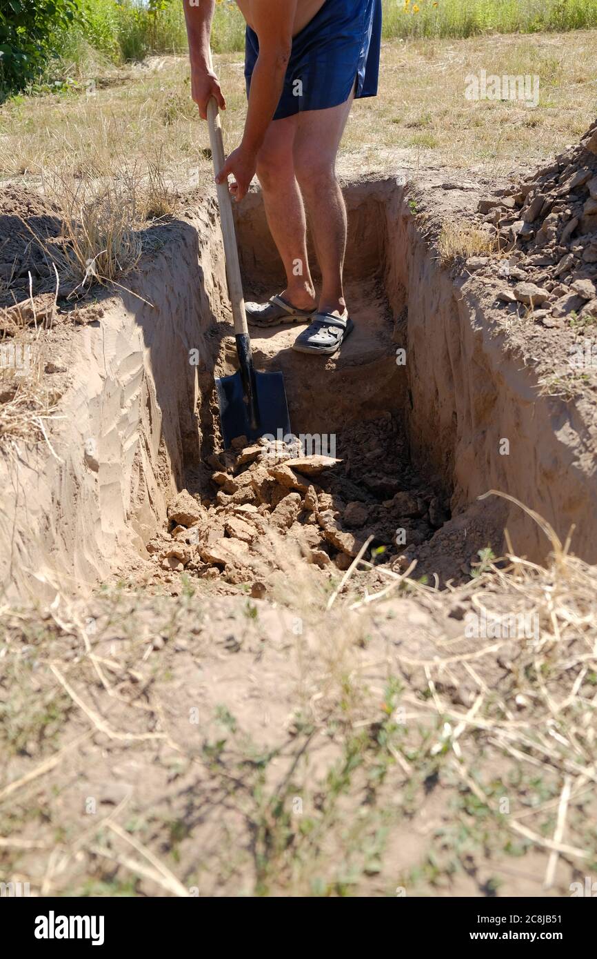 un uomo in pantaloncini blu scava un buco profondo con una pala. un uomo scava una tomba in un cimitero nel pomeriggio. Foto Stock