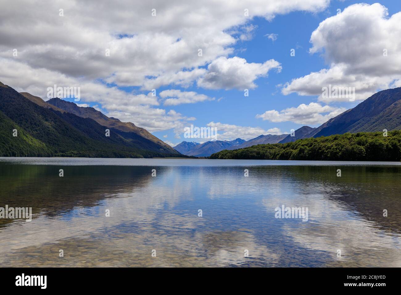 Le acque calme del Lago di Mavora Sud con montagne boscose lungo ogni lato e montagne in lontananza. Nuvole bianche e soffici su un cielo blu. Foto Stock