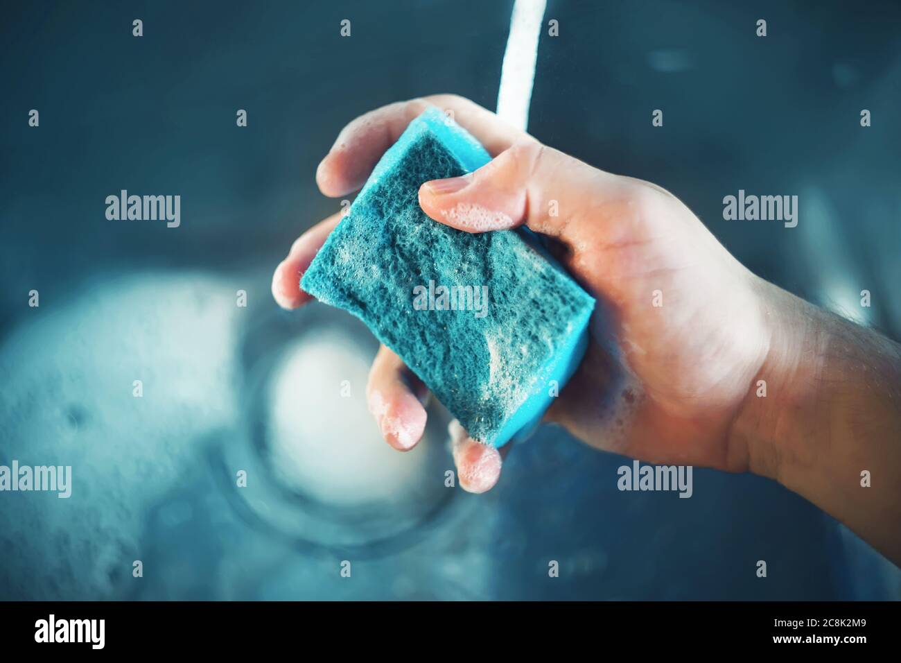 Un uomo tiene una spugna blu con schiuma in mano sopra il lavandino della cucina, dal cui rubinetto scorre l'acqua. Stava lavando i piatti. CH. Casa Foto Stock