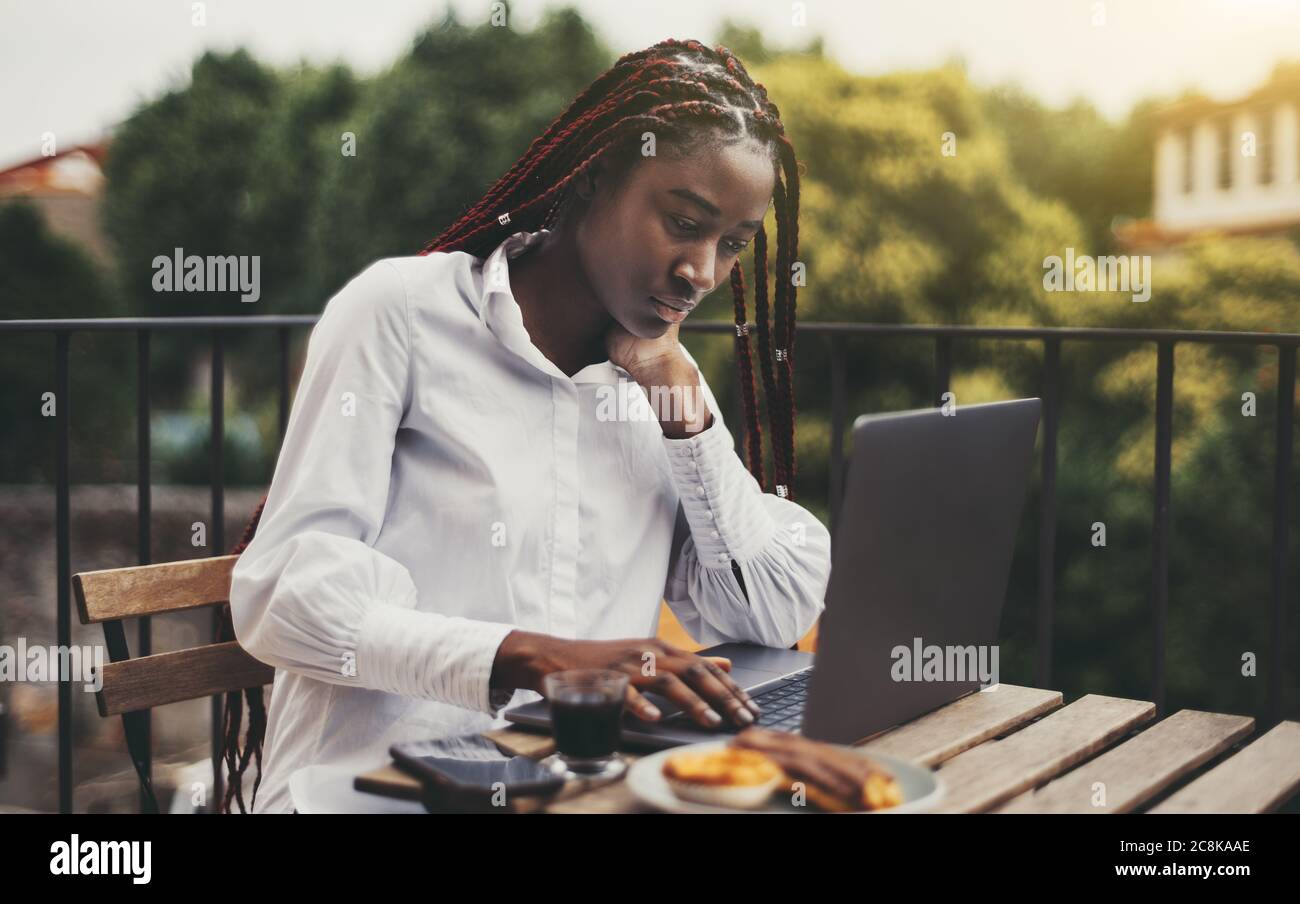 Una giovane e ravicante freelancer nera sta lavorando a distanza con il suo progetto utilizzando un computer portatile mentre si siede al tavolo con una tazza di caffè Foto Stock