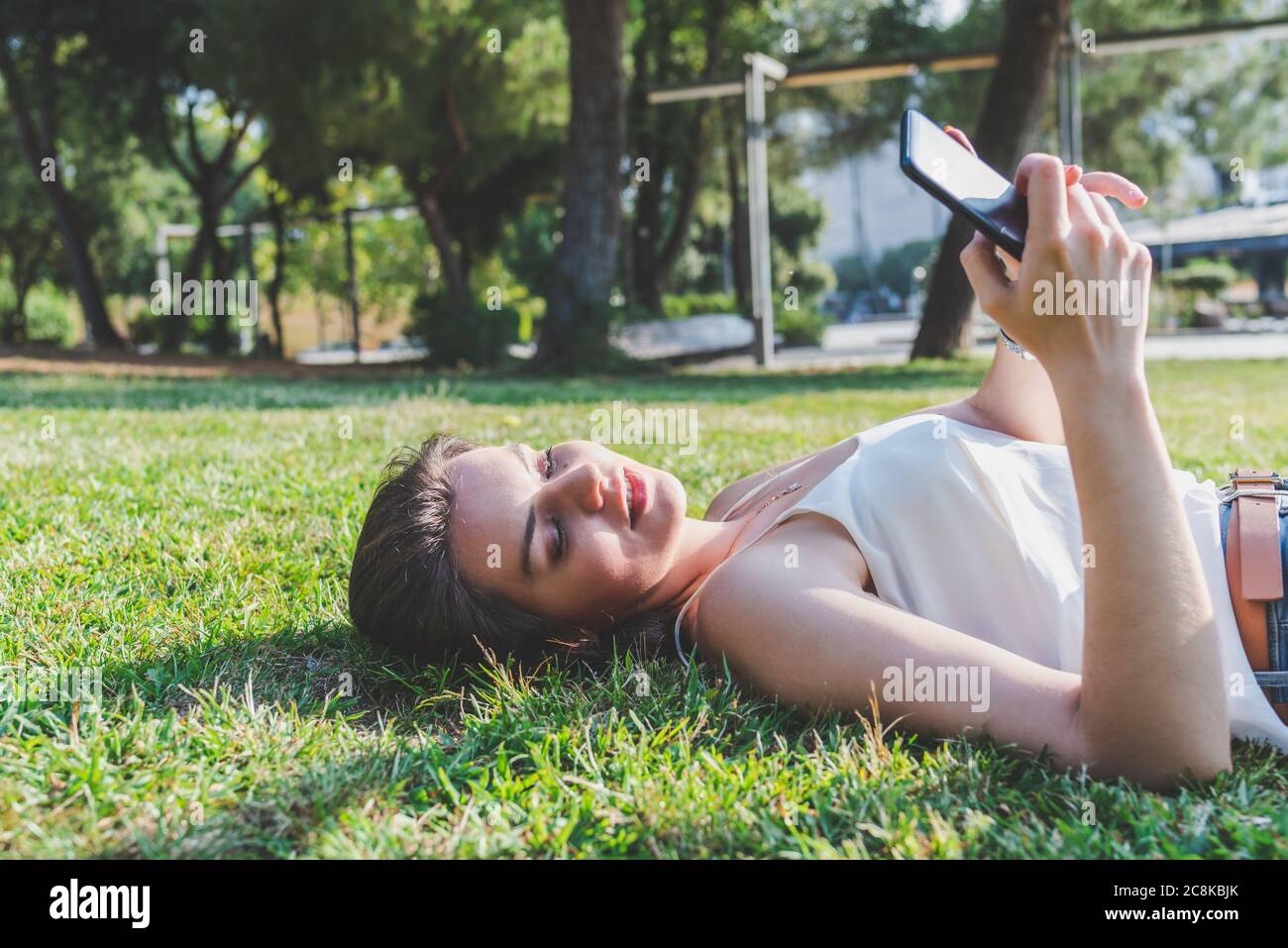 bella giovane donna caucasica che si stende sull'erba di un parco urbano e usa un telefono Foto Stock