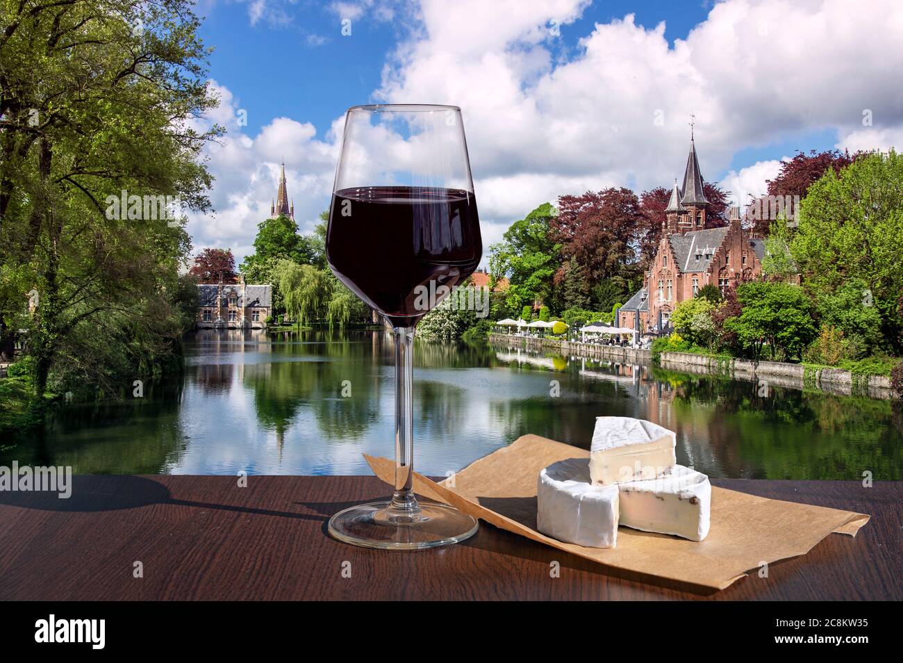 Bicchiere di vino rosso con vista sul tranquillo spazio verde pubblico con il lago Minnewater e il piccolo castello di Bruges durante la giornata di sole in primavera, Belgio Foto Stock