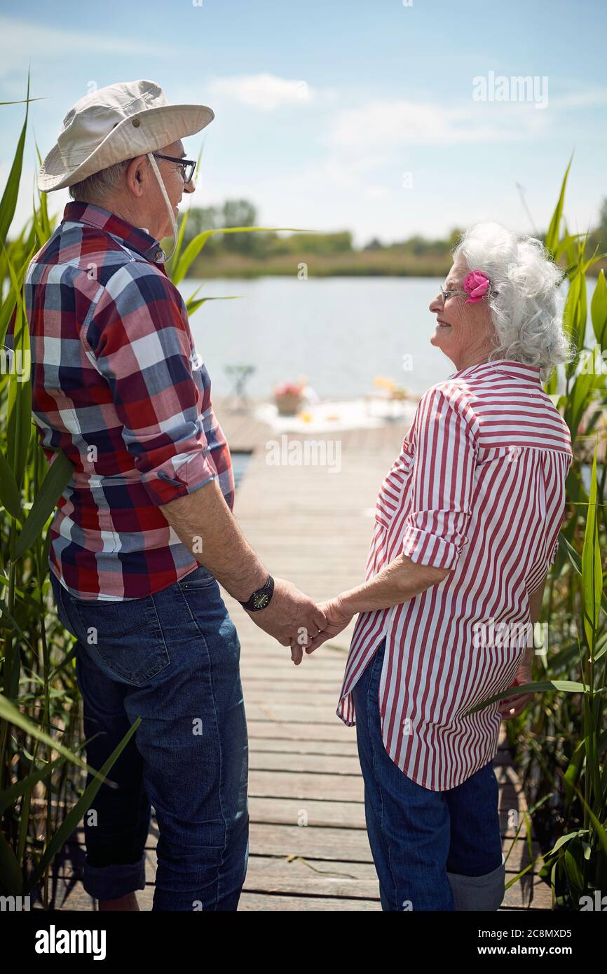 primo piano di carina coppia caucasica senior che tiene le mani, guardandosi l'un l'altro, sorridendo, in piedi presso il lago Foto Stock