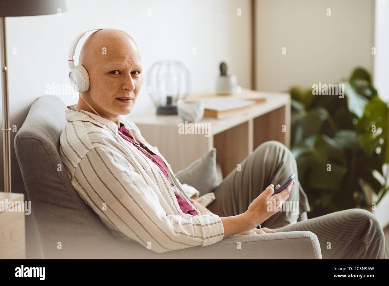 Ritratto con vista laterale minima di una donna adulta calva che indossa le cuffie e guarda la fotocamera mentre ascolti la musica tramite smartphone in un ambiente accogliente, Foto Stock