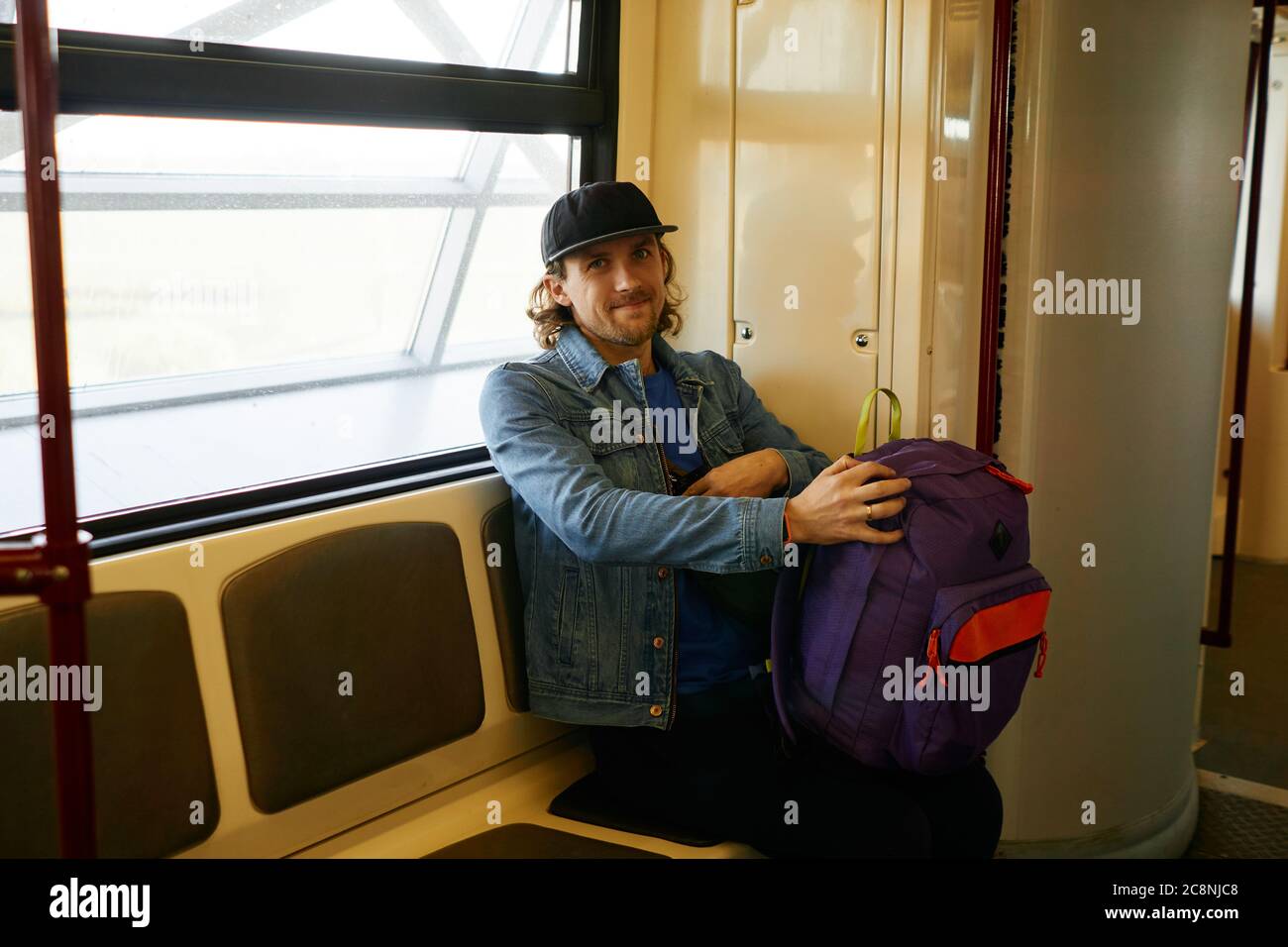 Felice giovane viaggiatore con bearded cappello da baseball e giacca jeans con  zaino viola seduto all'interno del treno della metropolitana nel paese  europeo. Turismo, viaggi Foto stock - Alamy