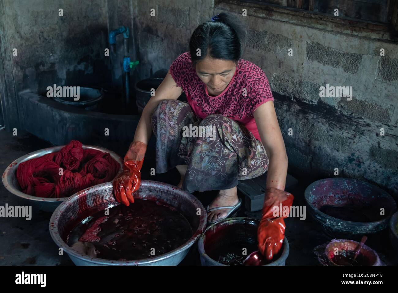 Donna asiatica tessuto rosso morente a mano utilizzato nella produzione di accappatoi per i monaci buddisti. Femmina accoccolato su ciotole di metallo rotonde in una stanza buia Foto Stock