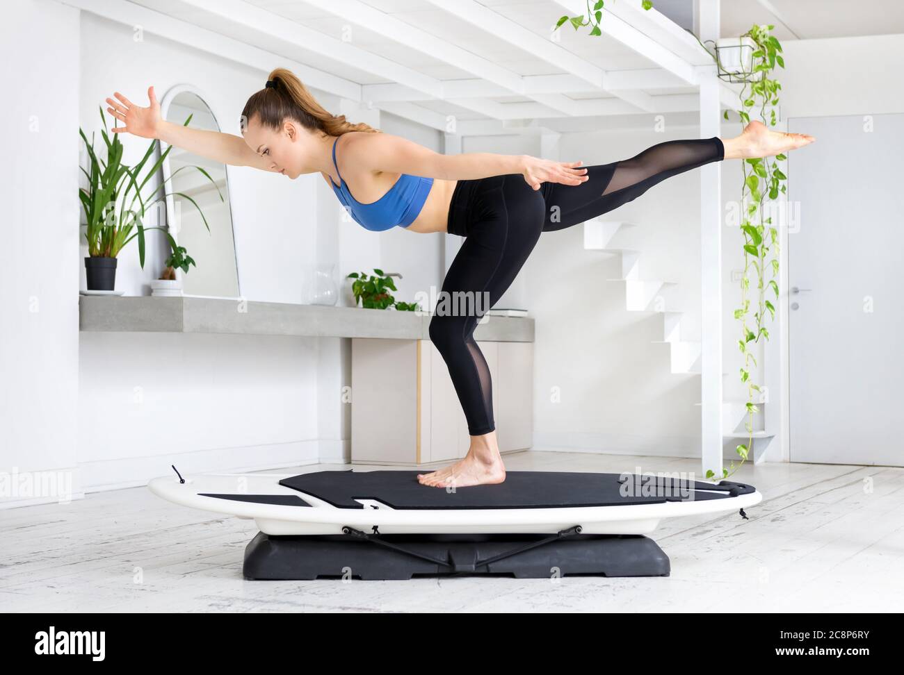 Giovane donna che fa una guida di yoga di surf posa equilibrare su una gamba con le braccia tese in una palestra chiave alta con piante verdi in un concetto sano Foto Stock