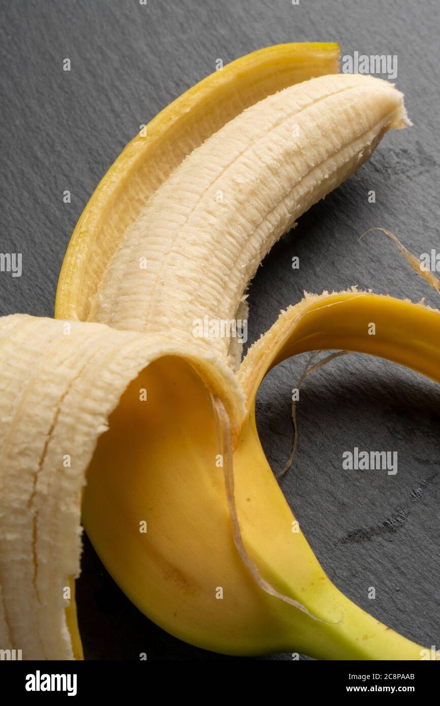 Banana gialla matura con bucce a metà buccia su una tavola scura di pietra grigia. Vista verticale. Concetto di dieta Foto Stock