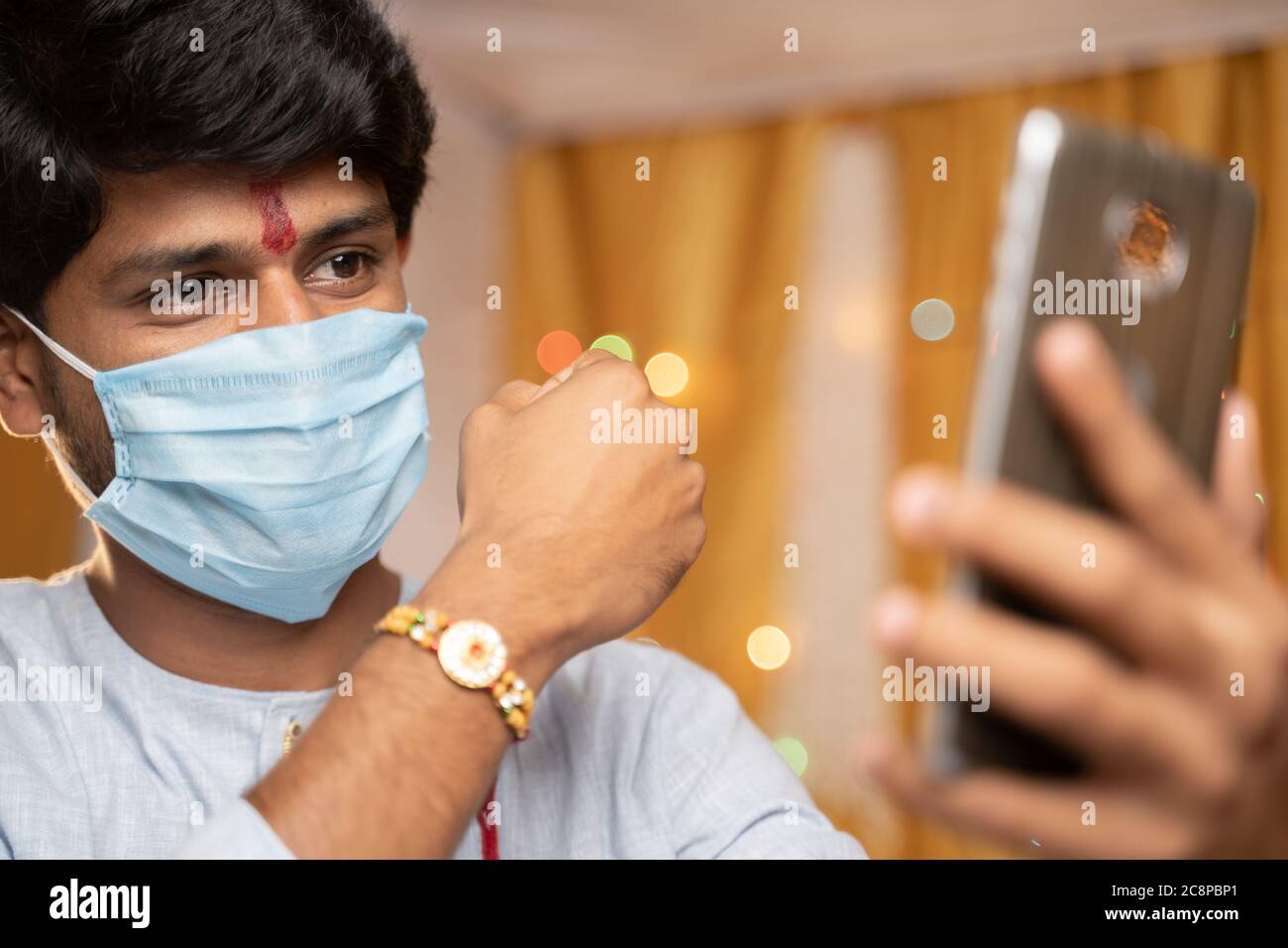 Uomo in maschera medica occupato sul telefono cellulare e mostrando Rakhi o RakshaBandhan a sorella o amici di famiglia alla cerimonia del festival durante il coronavirus o. Foto Stock