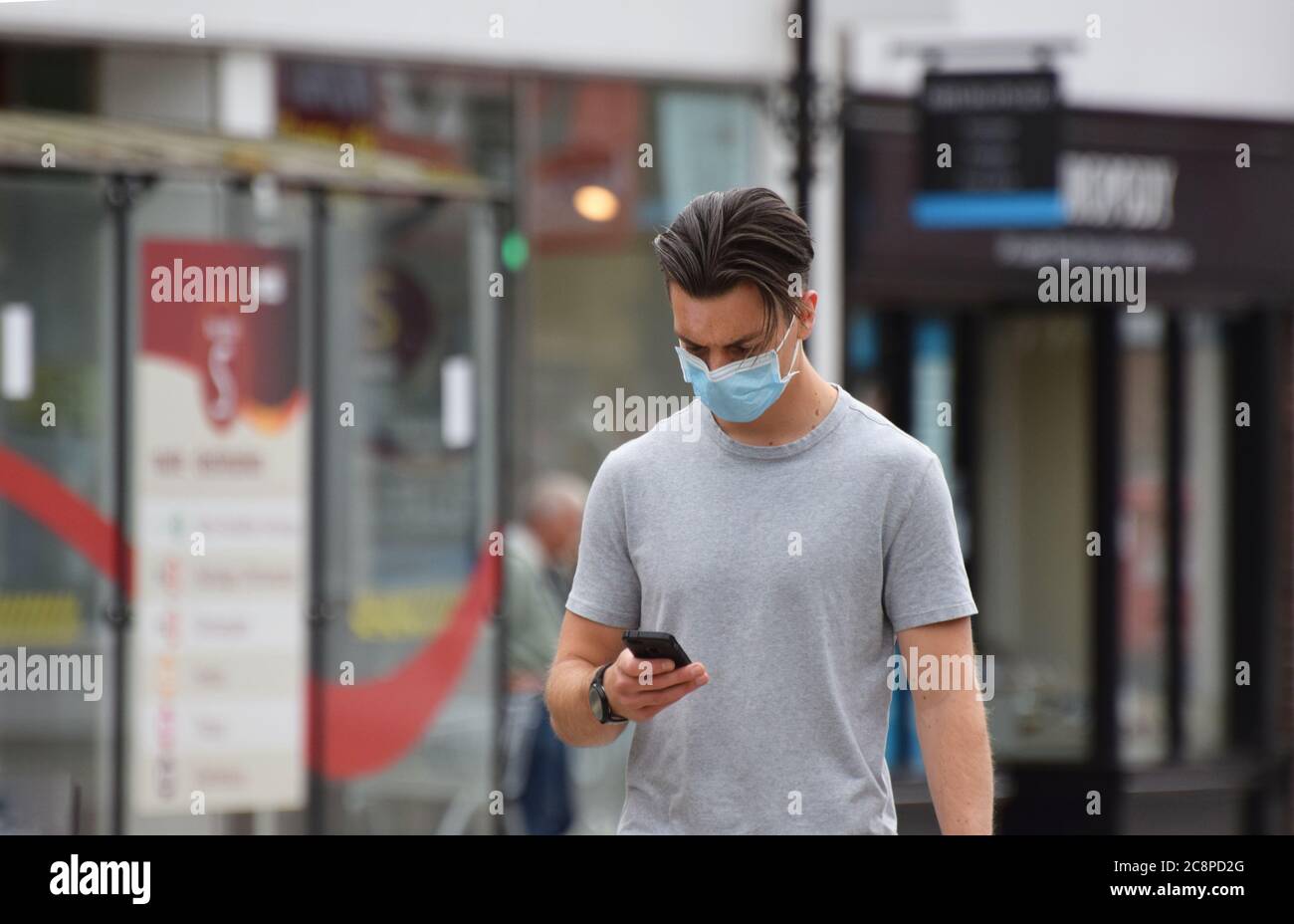 Un giovane inglese negli anni venti indossa una maschera facciale per strada mentre cammina e testi sul suo smartphone Foto Stock