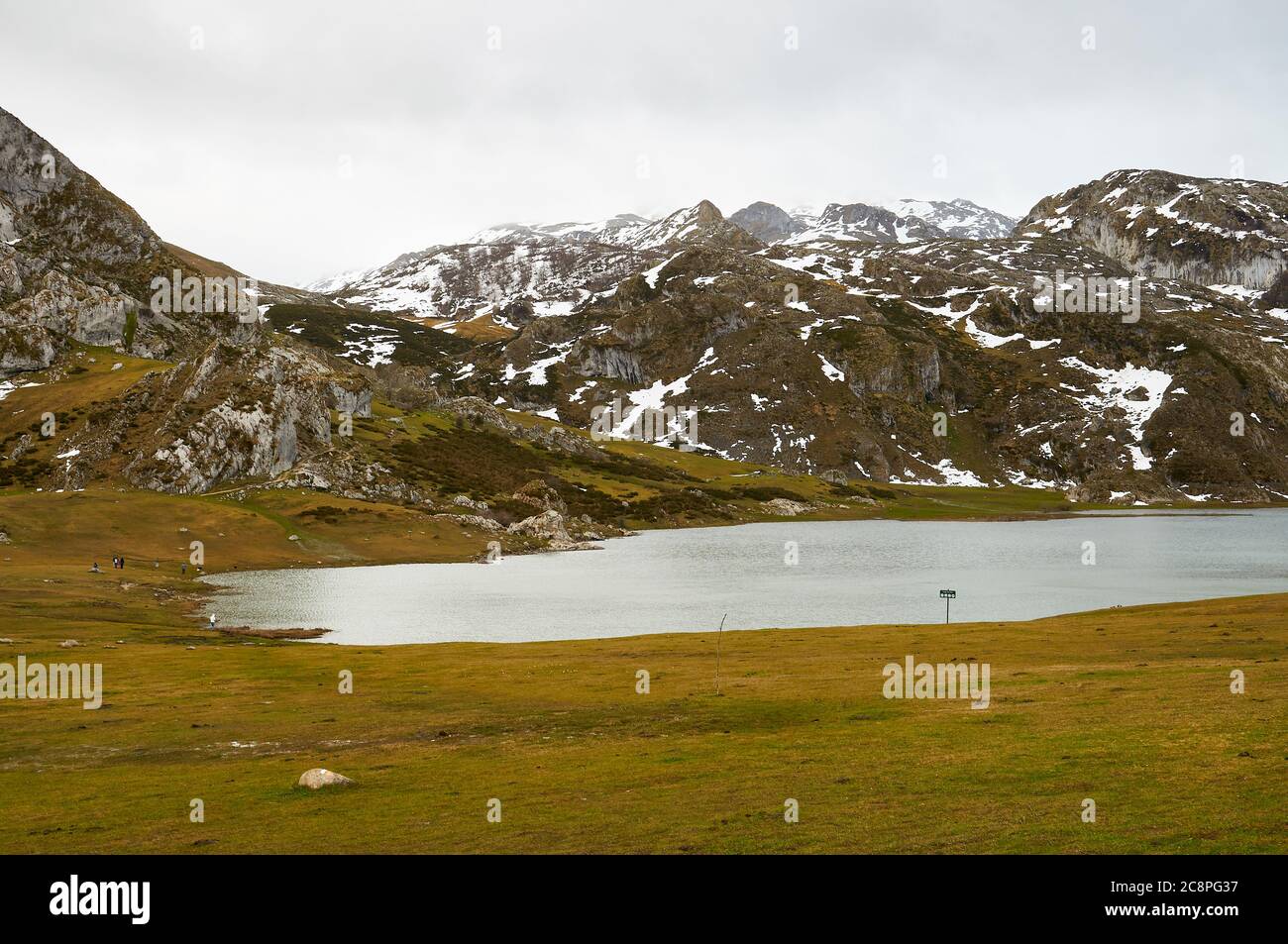 Escursionisti intorno al lago glaciale del Lago Ercina in una giornata nuvolosa con neve nelle cime vicine (Cangas de Onís, Parco Nazionale Picos de Europa, Asturie, Spagna) Foto Stock