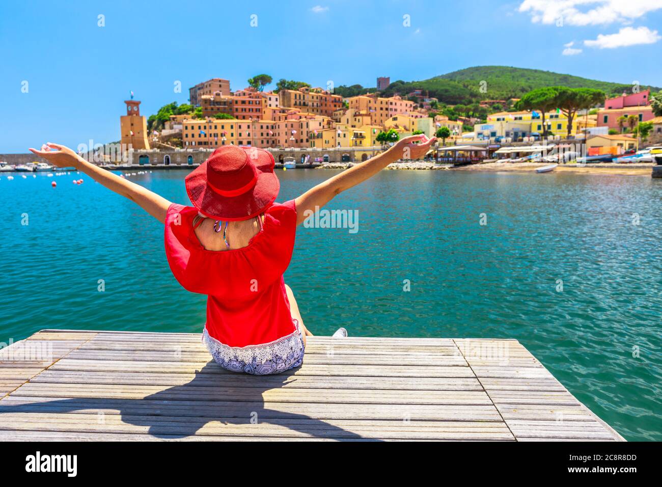 Felice donna seduta sopra il molo e guardando Rio Marina porto dell'isola d'Elba in tuta rossa. Ragazza turistica lifestyle in viaggio di vacanza in Italia, Europa Foto Stock