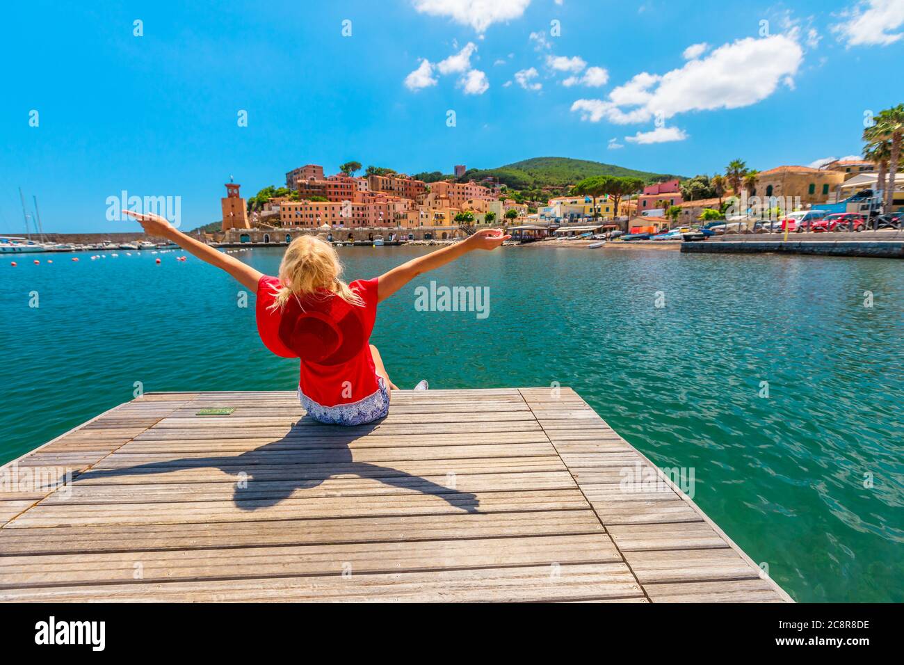 Donna turistica seduta sul molo e guardando il porto di Rio Marina dell'isola d'Elba in costume rosso. Ragazza turistica lifestyle in viaggio di vacanza in Italia Foto Stock
