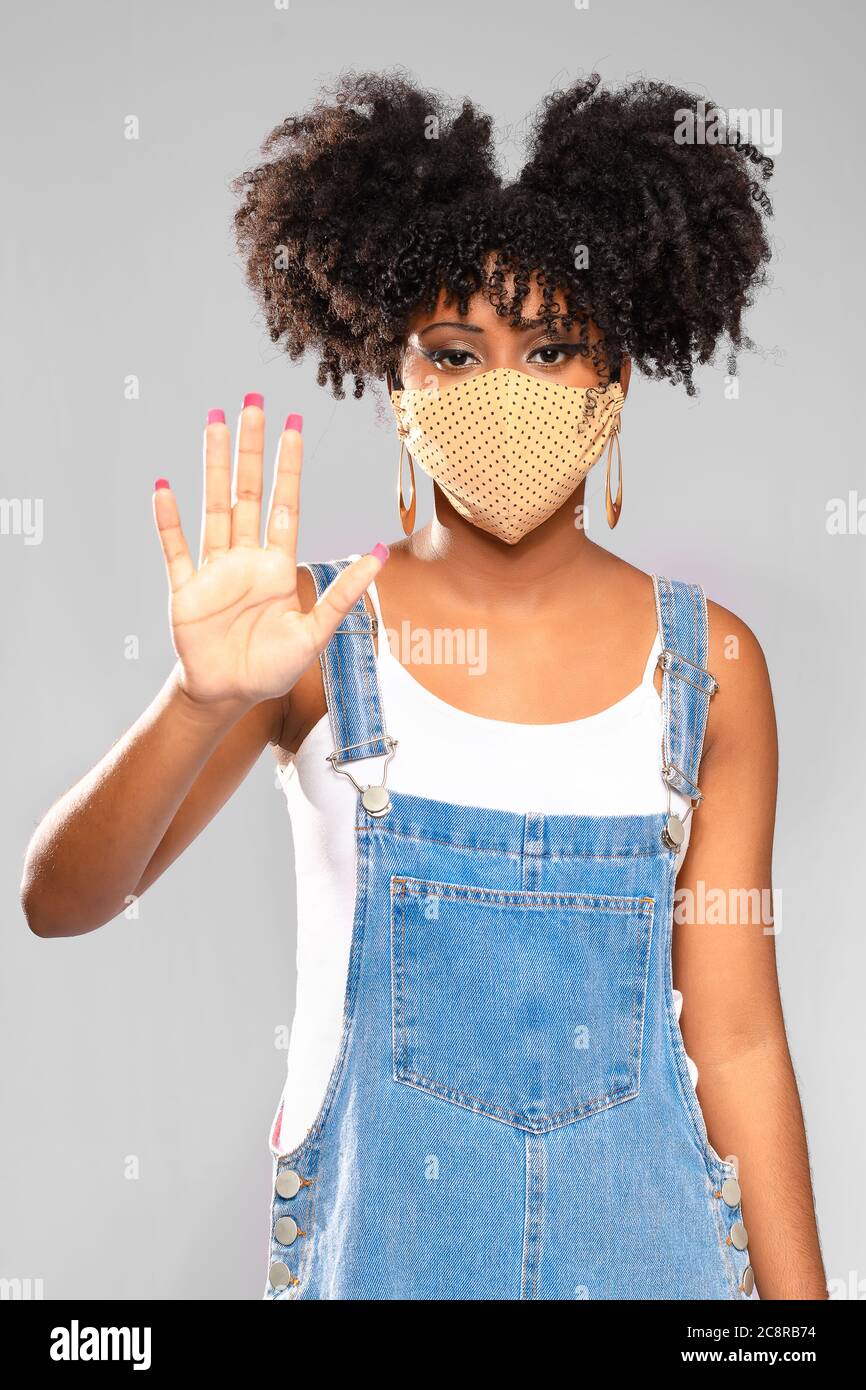 Afro-brasiliano teenage modello gesti con una mano di stop e punta all'uso di una maschera protettiva contro INVITE-19 Foto Stock
