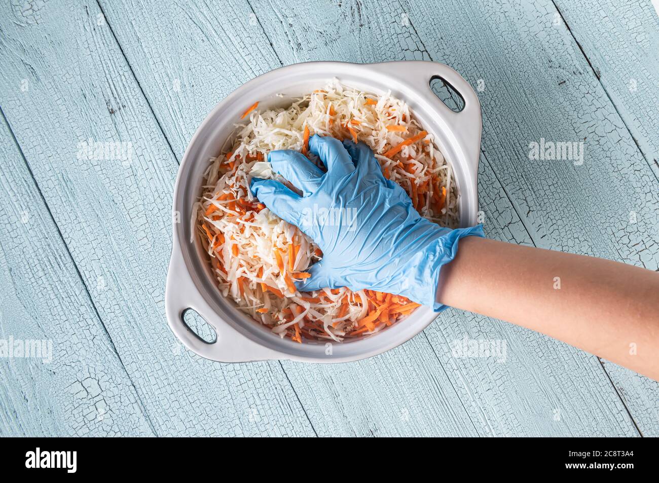 Mano di adolescente in guanto blu spremitura cavolo grattugiato e carota in padella su tavola di legno blu. Piatto di cottura per insalata di cavolo fermentato e sala Foto Stock