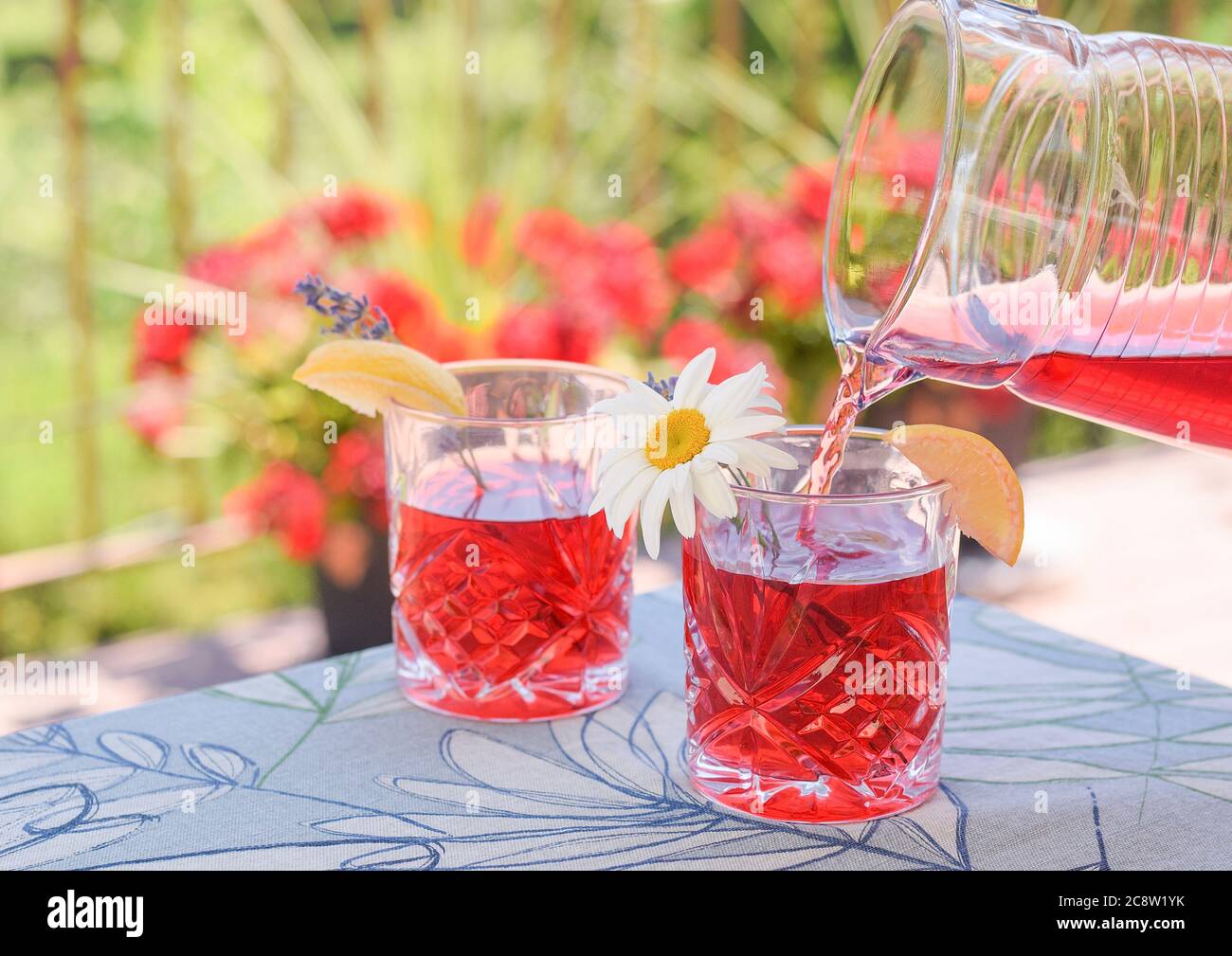 Caraffa versando una rinfrescante bevanda a base di ghiaccio rosso in bicchieri con fiori e limone Foto Stock