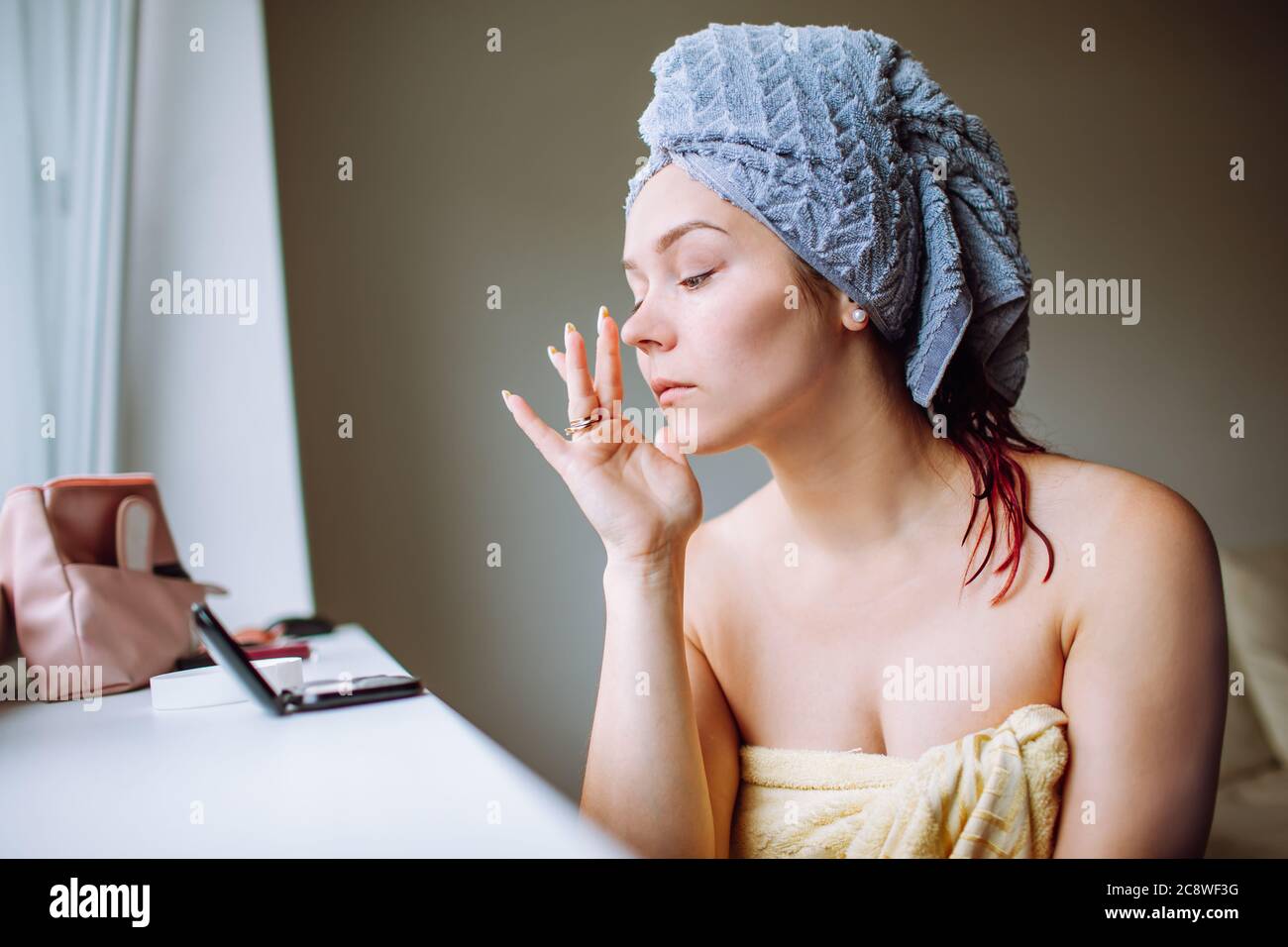 Bella giovane donna con asciugamano blu sulla testa applicando crema anti-rughe al viso mentre si guarda al piccolo specchio Foto Stock