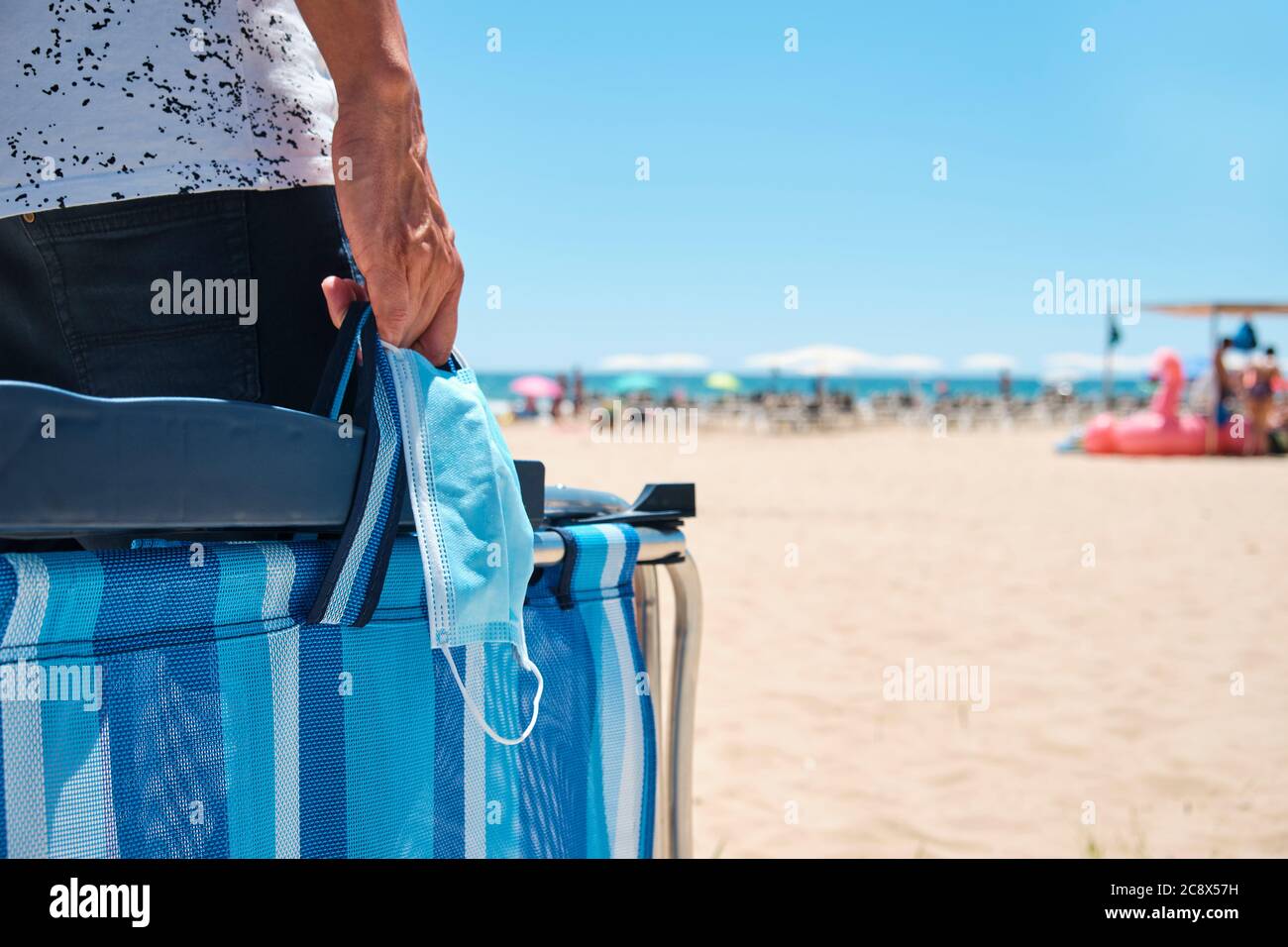 primo piano di un uomo caucasico, indossando pantaloncini, portando una maschera chirurgica e una sedia da spiaggia ripiegata nel suo cammino verso la spiaggia, con il mare sullo sfondo Foto Stock