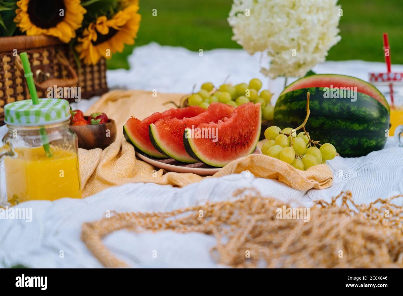 Estate picnic concetto in giornata di sole con anguria, frutta, bouquet di  idrangea e fiori di girasole. Cestino da picnic sull'erba con cibo e  bevanda estiva rinfrescante su coperta bianca in maglia.